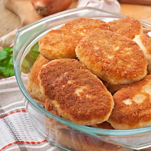 A glass bowl filled with golden-brown chicken cutlets, placed on a checkered kitchen towel with garlic, onions, and parsley in the background.