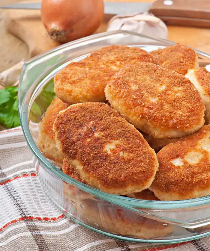 A glass bowl filled with golden-brown chicken cutlets, placed on a checkered kitchen towel with garlic, onions, and parsley in the background.