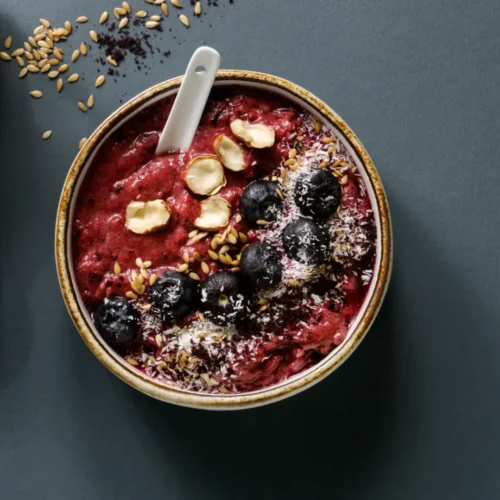 A dark berry smoothie bowl topped with blueberries, shredded coconut, flaxseeds, and sliced nuts, served in a rustic ceramic bowl with a white spoon on a dark background.