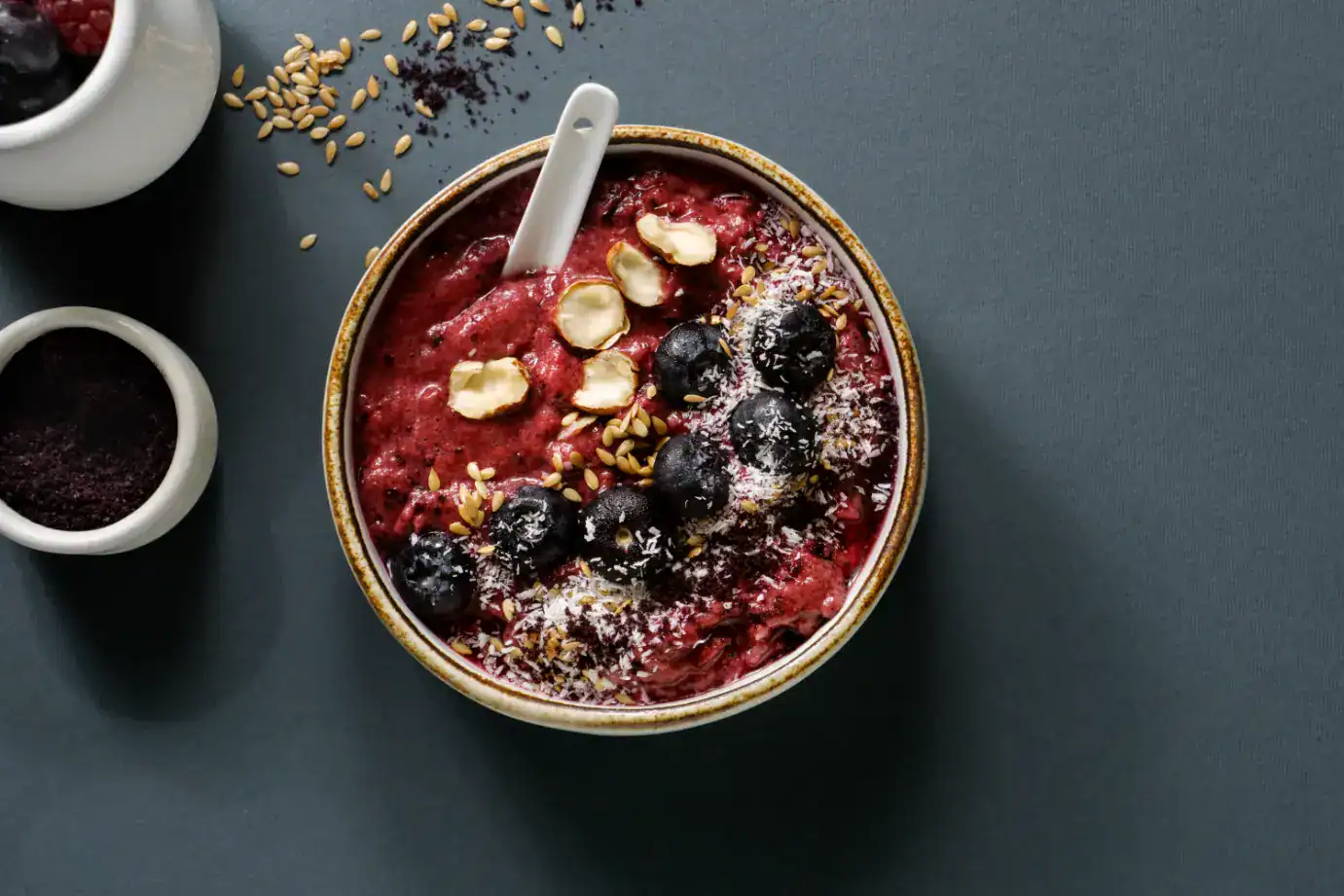 A dark berry smoothie bowl topped with blueberries, shredded coconut, flaxseeds, and sliced nuts, served in a rustic ceramic bowl with a white spoon on a dark background.