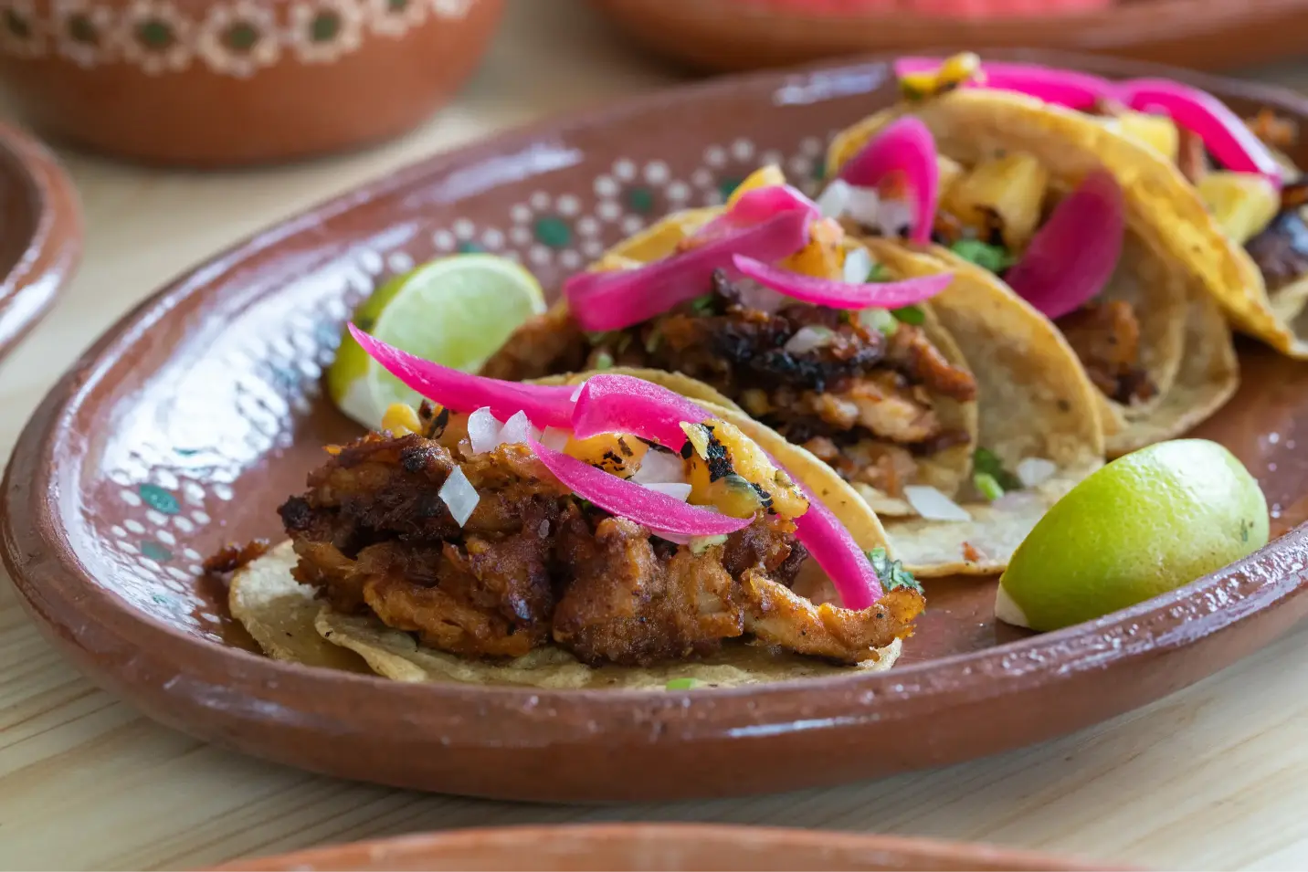 Close-up of traditional Mexican tacos al pastor served on a decorative plate with lime wedges and vibrant toppings like pickled onions and fresh cilantro.