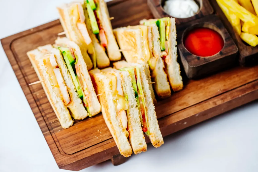 A wooden platter featuring classic club sandwiches layered with toasted bread, fresh vegetables, and melted cheese, served alongside French fries and dipping sauces.