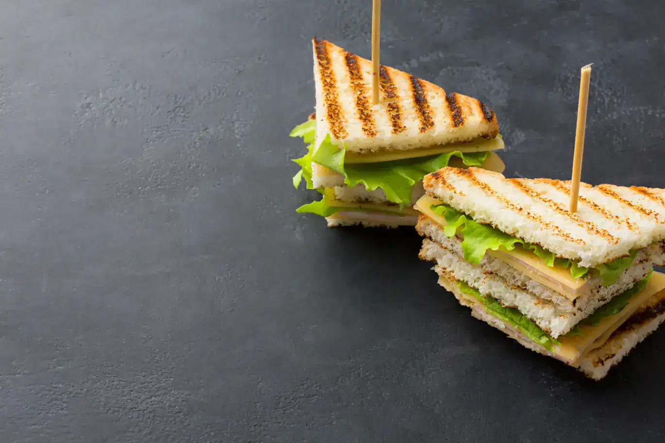 Two neatly arranged triangular club sandwiches with layers of toasted bread, lettuce, cheese, and deli meat, secured with toothpicks on a dark slate background.