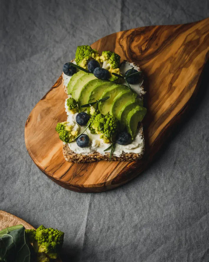 A slice of whole grain toast topped with creamy spread, sliced avocado, blueberries, and Romanesco broccoli, served on a wooden cutting board.