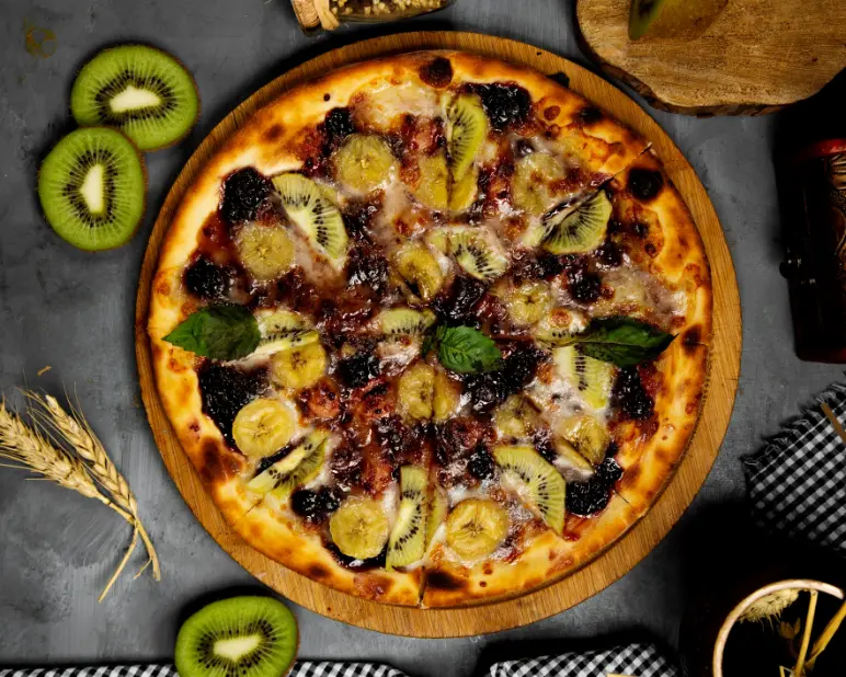 A beautifully baked fruit pizza topped with sliced kiwi, banana, and dark berries, garnished with fresh basil leaves, served on a wooden board.