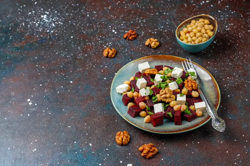 A beautifully plated beet and chickpea salad with feta cheese, walnuts, and fresh parsley, served on a ceramic plate with a side bowl of chickpeas and scattered walnuts on a rustic dark background.