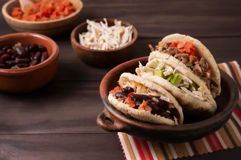 A selection of traditional Mexican tacos filled with shredded meat, avocado, beans, and fresh toppings, served in a rustic clay dish alongside bowls of additional ingredients.