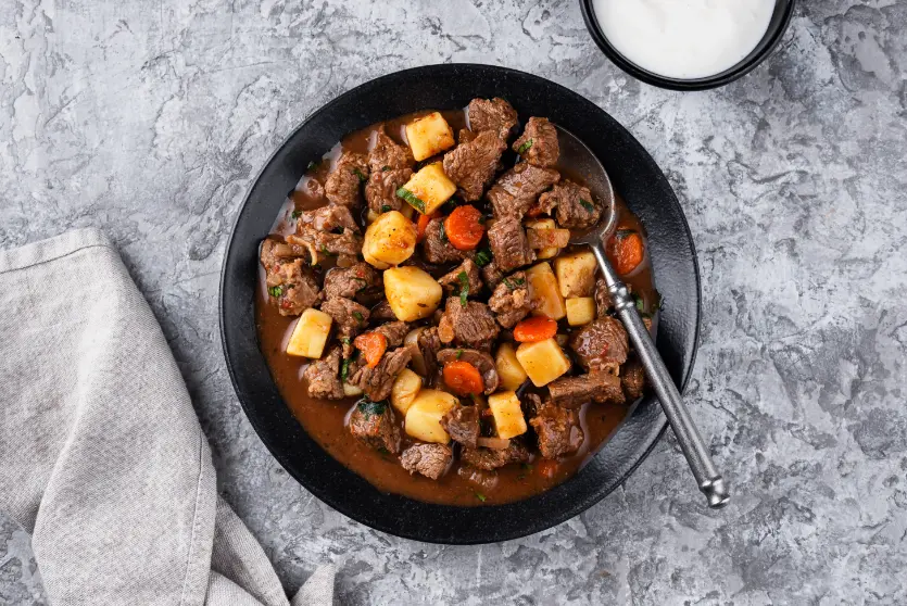 A hearty beef goulash served in a black bowl, filled with tender beef chunks, carrots, and potatoes in a rich, savory broth, accompanied by a spoon.