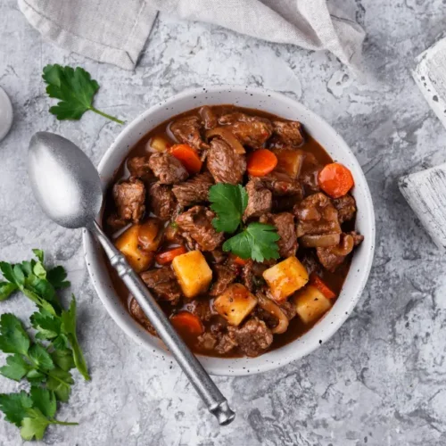 A bowl of hearty beef goulash filled with tender beef chunks, carrots, potatoes, and rich broth, garnished with fresh parsley, served with crusty bread.