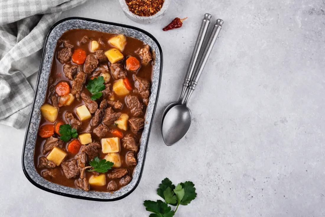 A rectangular dish filled with hearty beef goulash, featuring tender beef chunks, potatoes, carrots, and a rich brown sauce, garnished with fresh parsley.