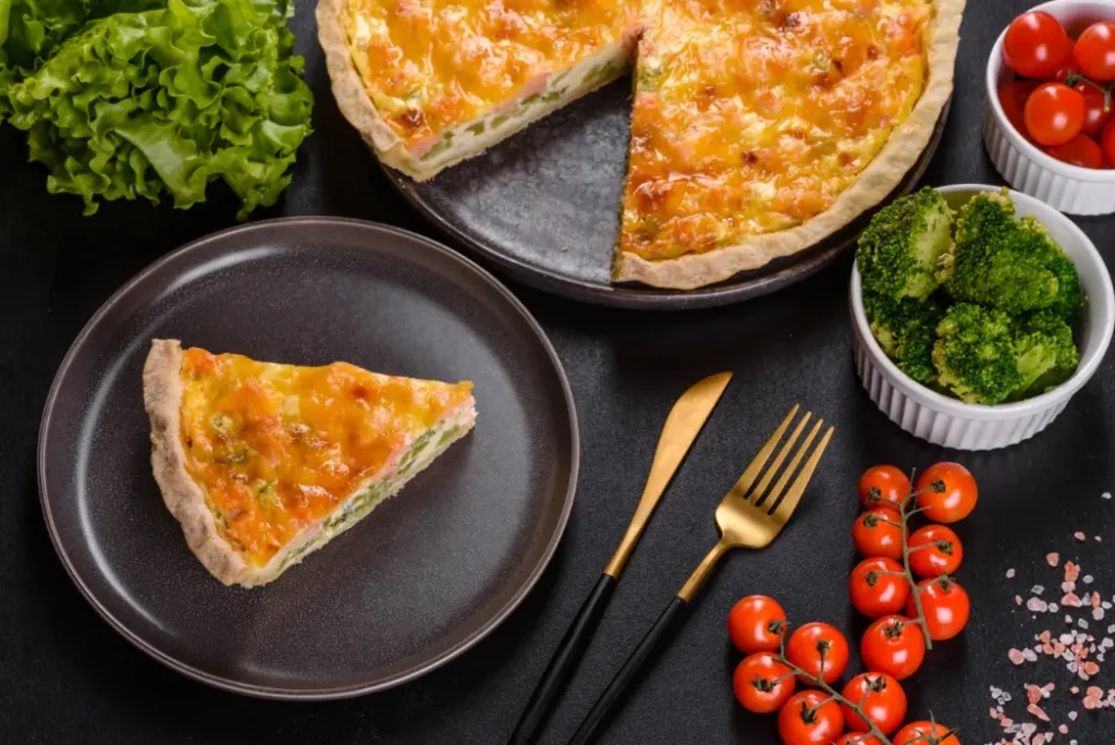 A golden-brown veggie quiche with a flaky crust, served on a dark plate with fresh lettuce, cherry tomatoes, and steamed broccoli on the side.