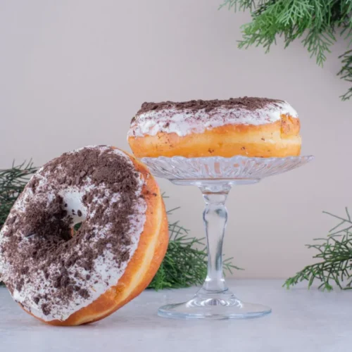 Two homemade doughnuts with a fluffy texture, coated in white glaze and sprinkled with crushed chocolate cookies, displayed on a glass stand.