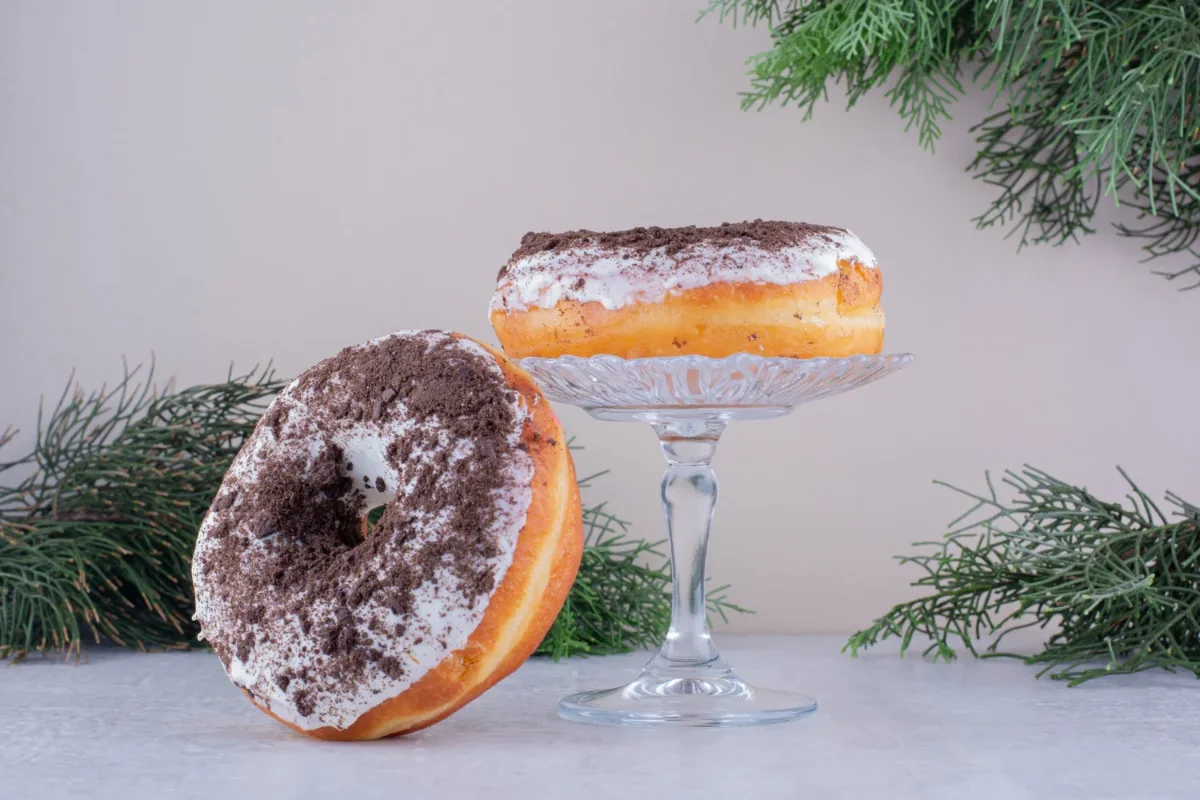 Two homemade doughnuts with a fluffy texture, coated in white glaze and sprinkled with crushed chocolate cookies, displayed on a glass stand.