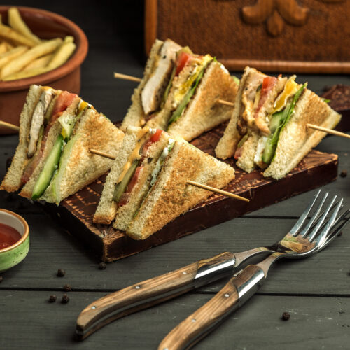 Four neatly sliced club sandwich triangles with layers of toasted bread, fresh vegetables, grilled chicken, and cheese, secured with bamboo skewers on a dark wooden platter, surrounded by condiments and cherry tomatoes.