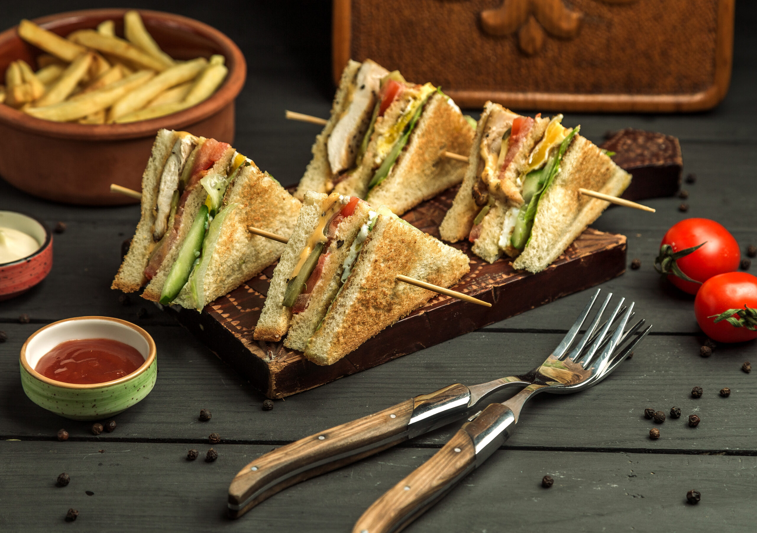 Four neatly sliced club sandwich triangles with layers of toasted bread, fresh vegetables, grilled chicken, and cheese, secured with bamboo skewers on a dark wooden platter, surrounded by condiments and cherry tomatoes.