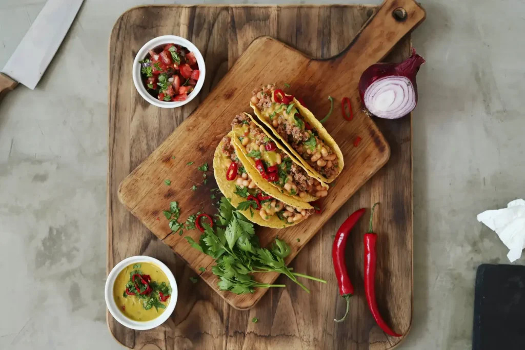 Overhead view of traditional Mexican tacos filled with beans, seasoned meat, and fresh toppings, served on a wooden board with bowls of salsa and garnishes.