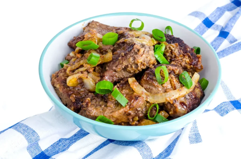 A bowl of perfectly cooked beef liver with caramelized onions, garnished with fresh green onions, placed on a blue and white kitchen towel.