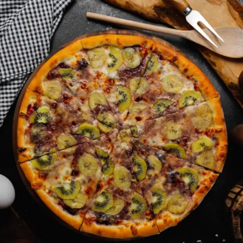 A freshly baked fruit pizza topped with sliced kiwi, banana, and dark berries on a golden crust, served on a wooden board.