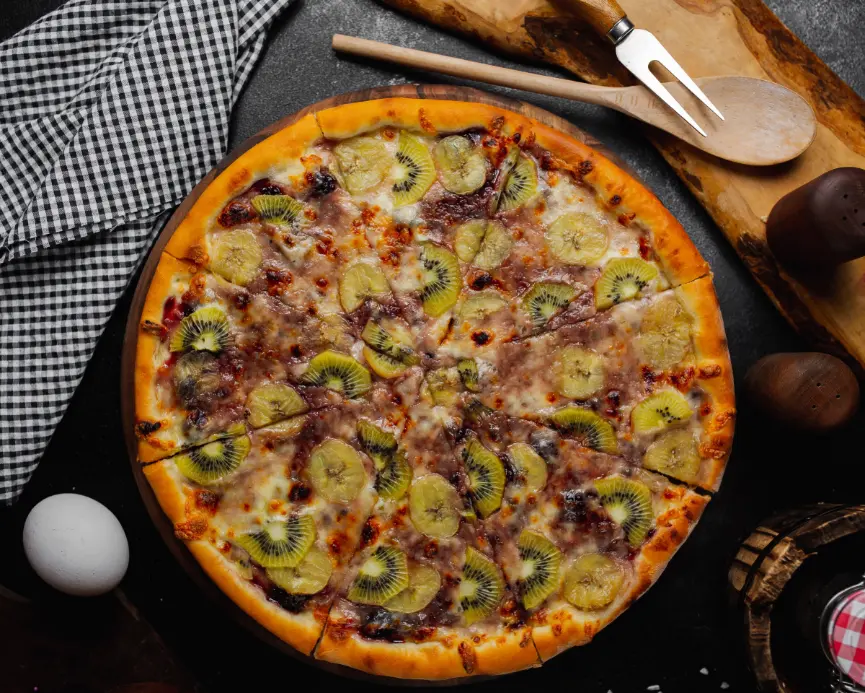 A freshly baked fruit pizza topped with sliced kiwi, banana, and dark berries on a golden crust, served on a wooden board.