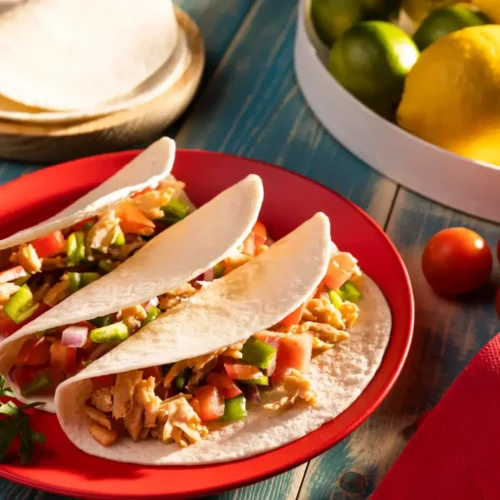 A vibrant plate of traditional Mexican tacos filled with shredded chicken, diced tomatoes, and bell peppers, served with fresh ingredients like lemons, limes, and cherry tomatoes.