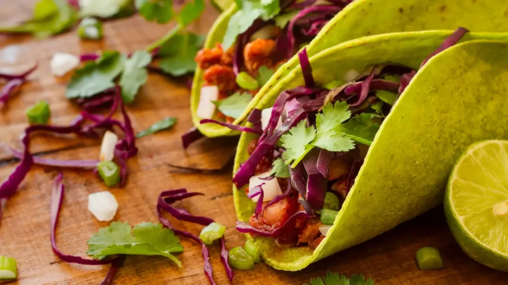 Close-up of vibrant Mexican tacos with green tortillas, red cabbage, fresh cilantro, and lime on a wooden table.