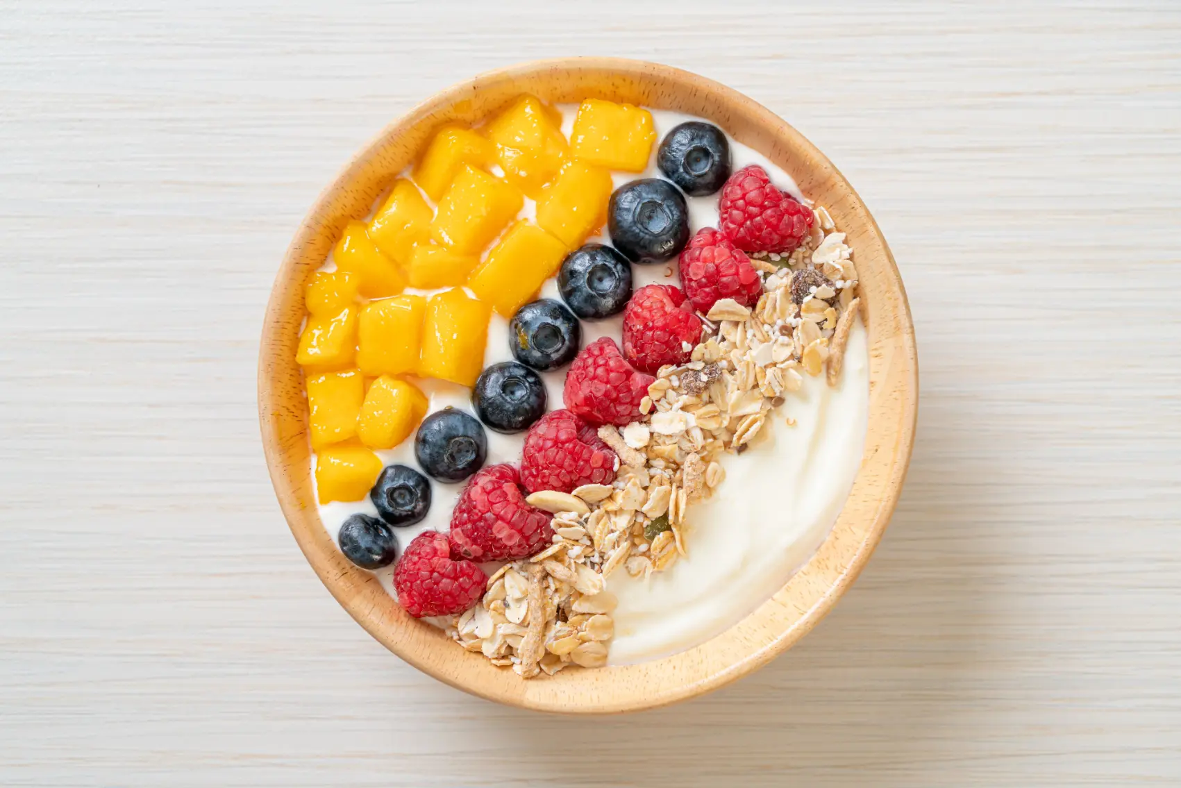 A creamy smoothie bowl topped with fresh mango chunks, blueberries, raspberries, granola, and chia seeds, served in a wooden bowl on a light-colored surface.