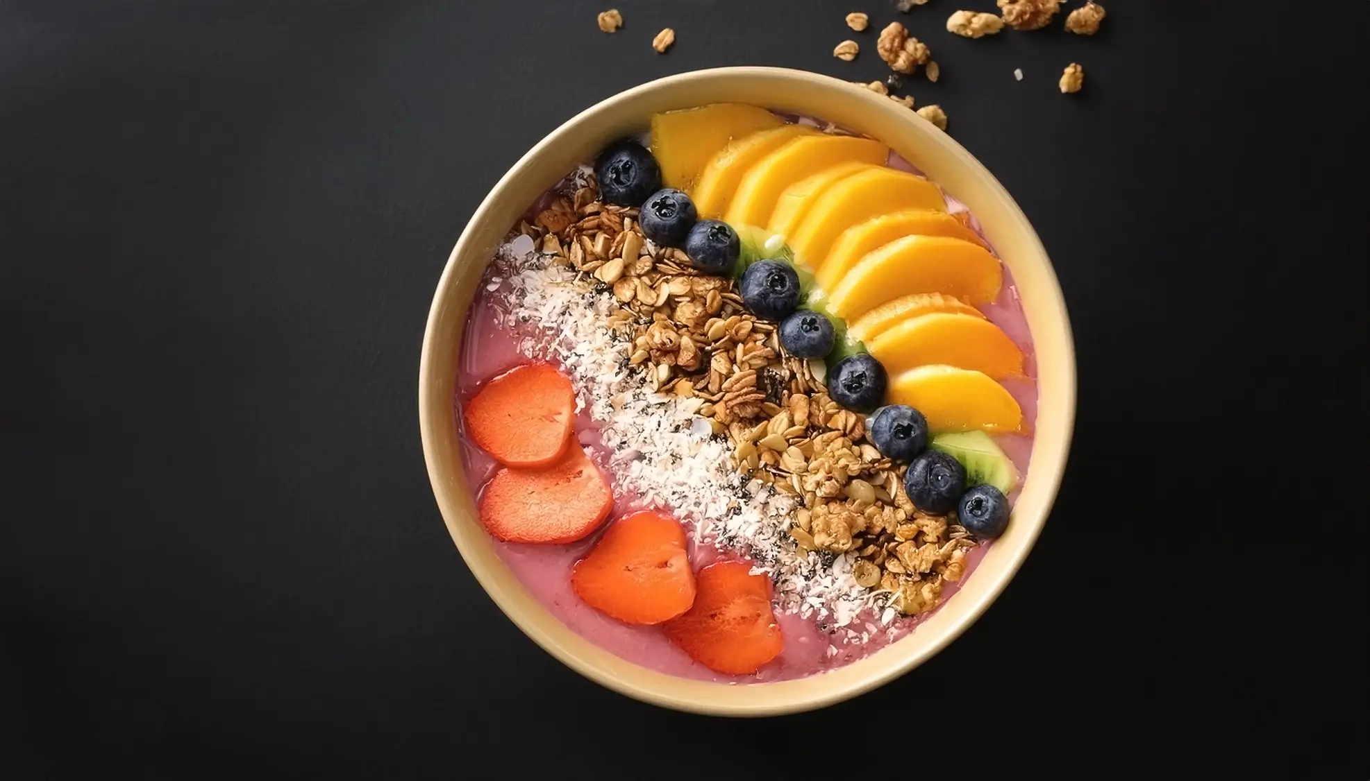 A vibrant smoothie bowl topped with fresh mango slices, blueberries, strawberries, granola, shredded coconut, and chia seeds, served in a yellow bowl on a dark background.