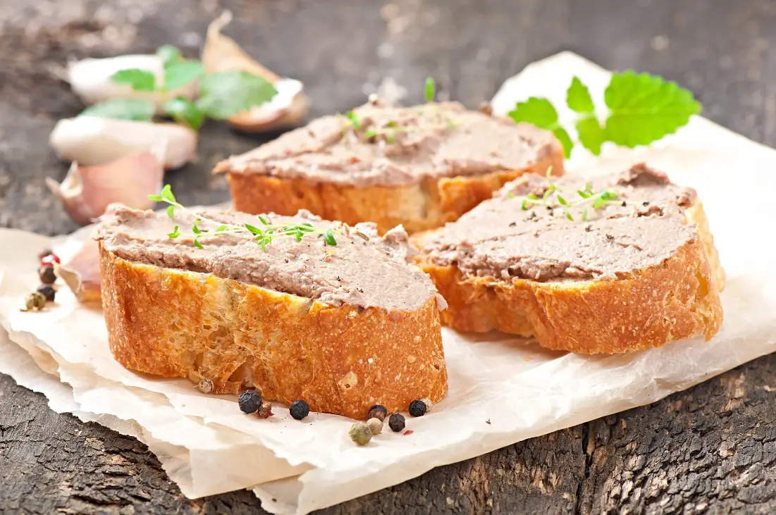 Slices of toasted baguette topped with creamy liver pâté and garnished with fresh herbs, served on parchment paper with whole peppercorns.