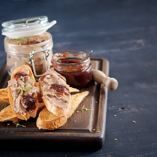 Slices of toasted baguette topped with smooth liver pâté and fruit preserves, served on a dark wooden board with jars of pâté and jam in the background.