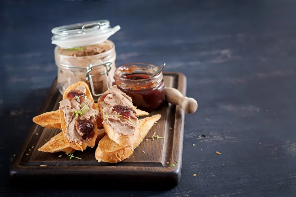 Slices of toasted baguette topped with smooth liver pâté and fruit preserves, served on a dark wooden board with jars of pâté and jam in the background.