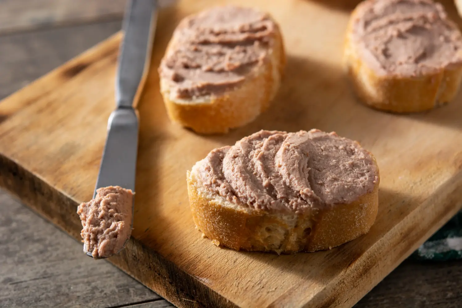 Slices of crusty baguette topped with creamy liver pâté, served on a wooden cutting board with a butter knife.