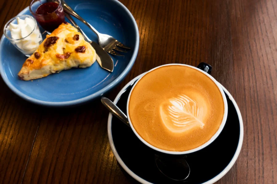 A freshly baked fruit scone served on a blue plate with whipped cream and jam, alongside a cup of latte with latte art on a wooden table.