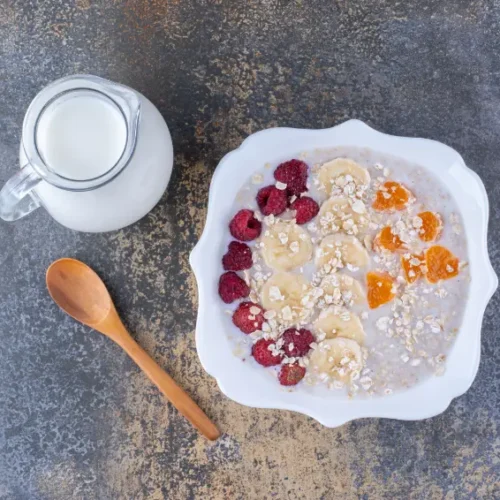 A bowl of creamy porridge topped with banana slices, raspberries, and orange pieces, served with a jug of milk and a wooden spoon on a dark textured surface.