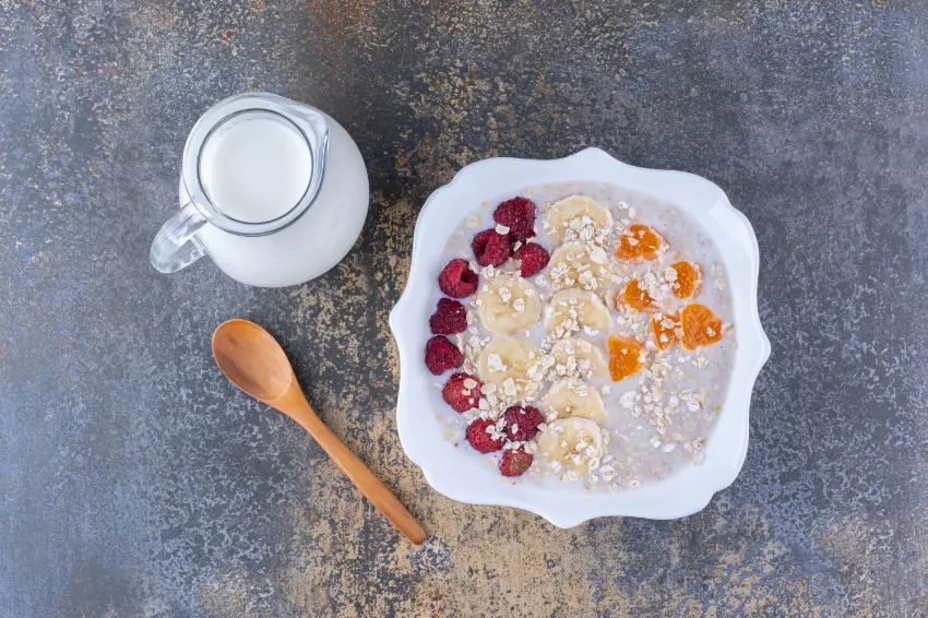 A bowl of creamy porridge topped with banana slices, raspberries, and orange pieces, served with a jug of milk and a wooden spoon on a dark textured surface.