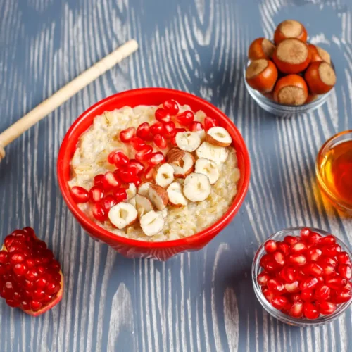 Two bowls of creamy oatmeal porridge topped with apple slices, pomegranate seeds, cinnamon, and honey, surrounded by spices and nuts on a rustic blue surface.