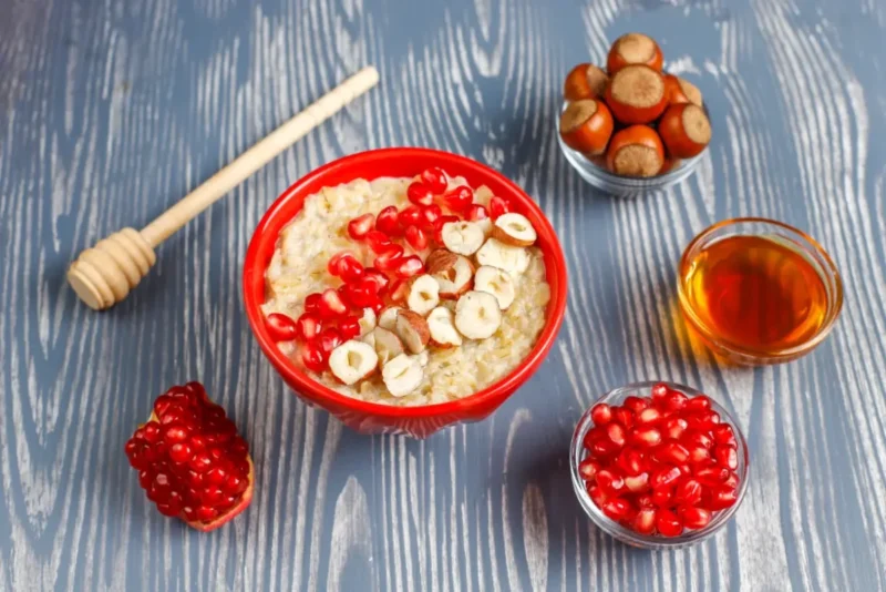 Two bowls of creamy oatmeal porridge topped with apple slices, pomegranate seeds, cinnamon, and honey, surrounded by spices and nuts on a rustic blue surface.