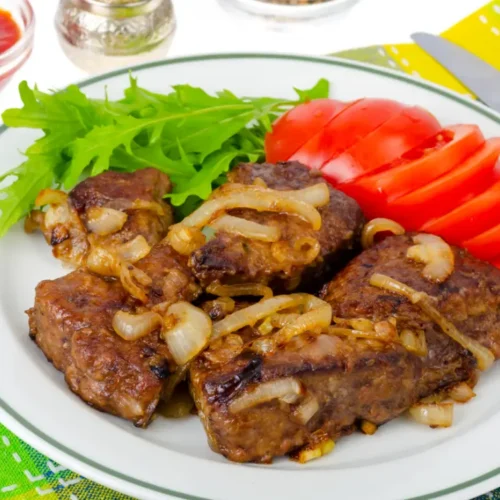 A plate of beef liver with caramelized onions, served alongside fresh lettuce and sliced tomatoes, on a colorful table setting.