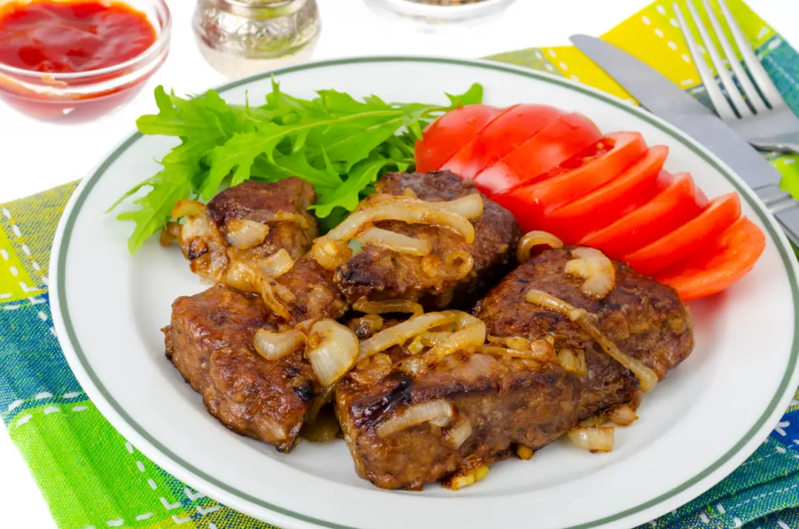 A plate of beef liver with caramelized onions, served alongside fresh lettuce and sliced tomatoes, on a colorful table setting.