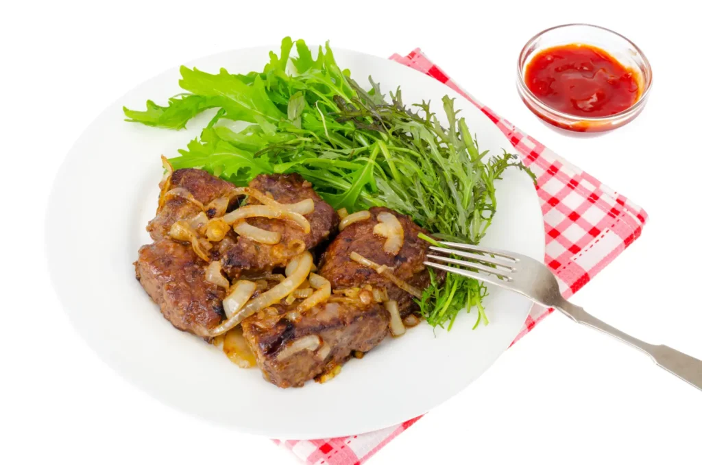 A plate of fried beef liver with caramelized onions, served with fresh green salad leaves, placed on a red and white checkered napkin.