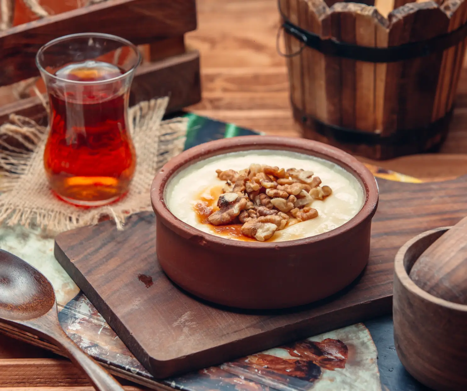 A rustic bowl of creamy porridge topped with walnuts and honey, served on a wooden board with a glass of tea in the background.