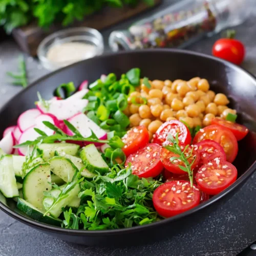 A vibrant chickpea salad in a black bowl with cherry tomatoes, sliced cucumbers, radishes, fresh greens, and herbs, topped with sesame seeds.