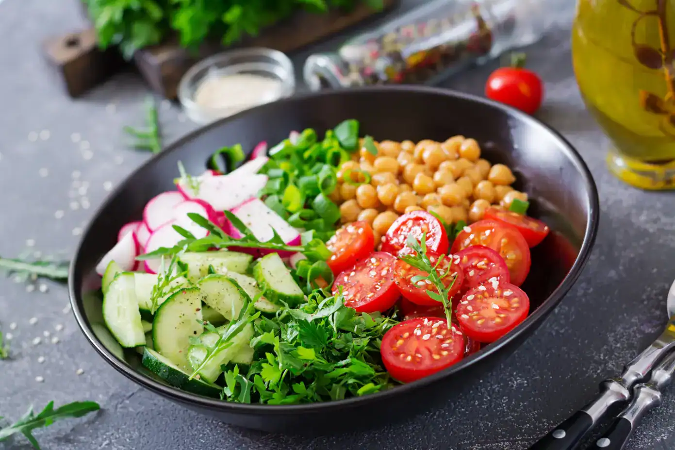 A vibrant chickpea salad in a black bowl with cherry tomatoes, sliced cucumbers, radishes, fresh greens, and herbs, topped with sesame seeds.