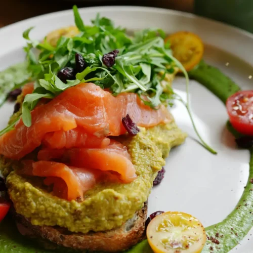 A gourmet avocado toast topped with smoked salmon, fresh arugula, and dried cranberries, served on a white plate with a green sauce and cherry tomatoes.