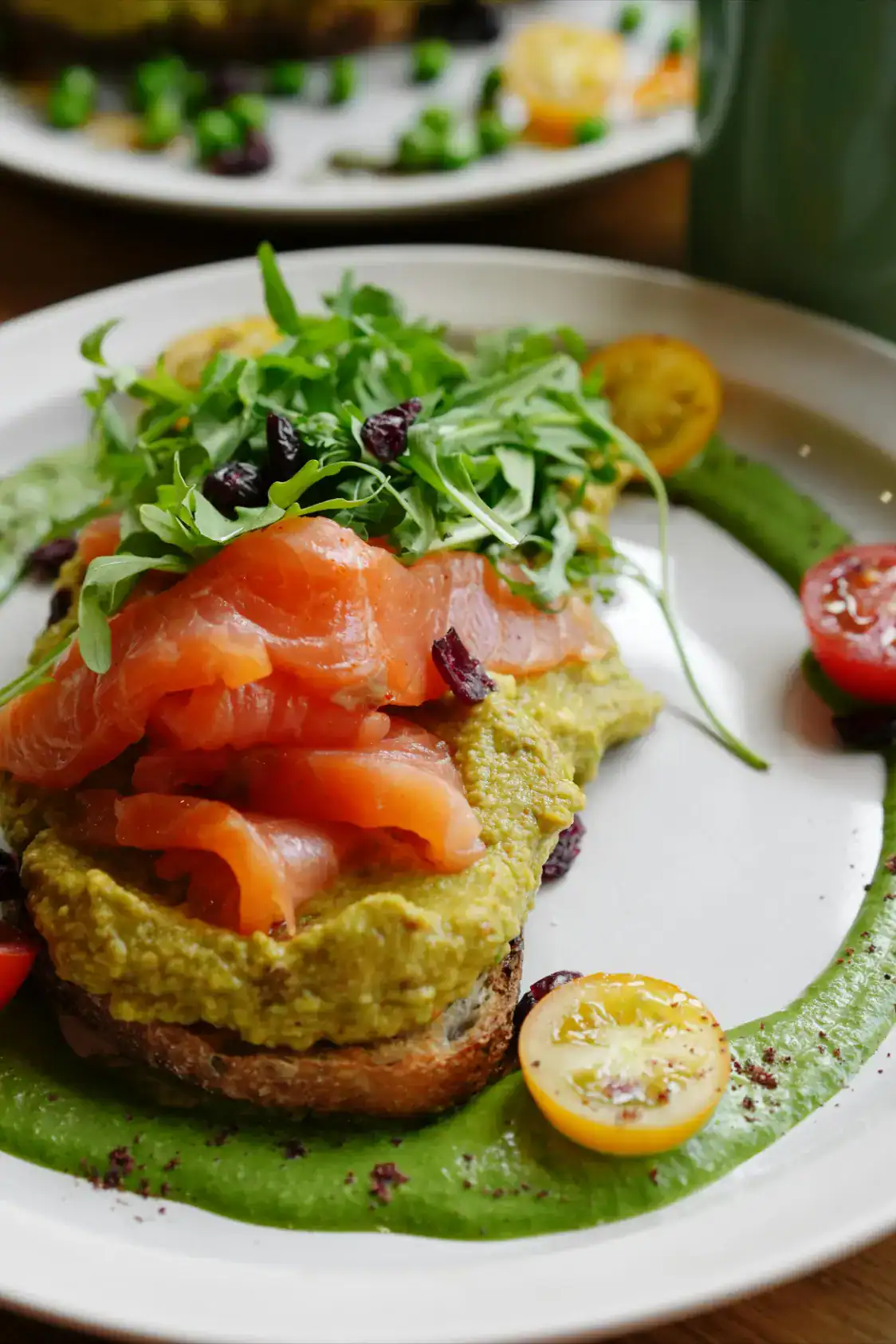 A gourmet avocado toast topped with smoked salmon, fresh arugula, and dried cranberries, served on a white plate with a green sauce and cherry tomatoes.