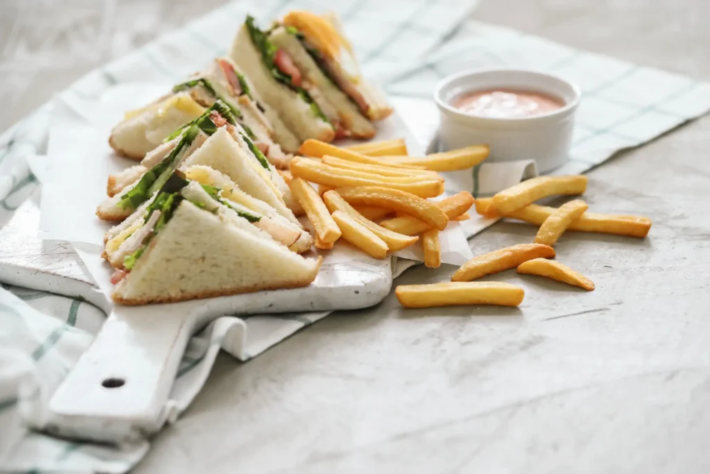 Triangular club sandwiches layered with lettuce, tomatoes, and meat, served with golden French fries and a side of creamy dipping sauce on a white serving board.