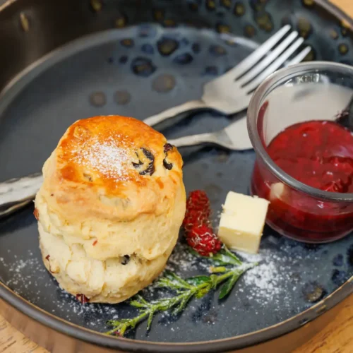 Traditional English scone with raisins, served with strawberry jam and butter on a black plate.