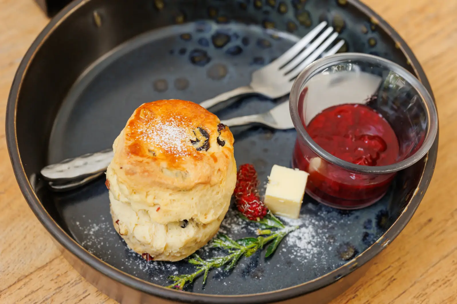 Traditional English scone with raisins, served with strawberry jam and butter on a black plate.