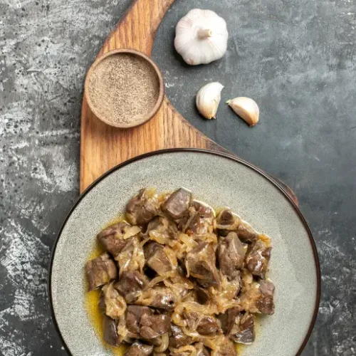 A plate of sautéed chicken livers with caramelized onions, served on a rustic wooden board with black pepper and garlic cloves on a textured grey background.