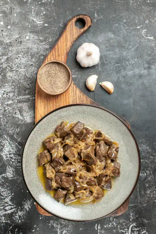 A plate of sautéed chicken livers with caramelized onions, served on a rustic wooden board with black pepper and garlic cloves on a textured grey background.