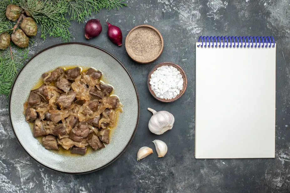 A plate of sautéed chicken livers with caramelized onions, served alongside fresh garlic, black pepper, sea salt, and an open notebook on a dark textured background.